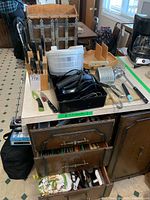 Photo of kitchen utensils including a wooden knife block with knives, hand mixer, salad spinner, cutting tool block, spice rack, metal grater, and assorted cutlery on a table