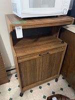 Microwave stand from a front angle with a white microwave on top, showing top shelf, open middle shelf, and closed cabinet doors with handles and caster wheels.