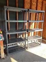 Two matching black plastic shelving units standing side by side in a garage setting with exposed wooden studs in the background and sunlight illuminating the right side.