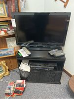 Full view of Panasonic 32-inch TV on black entertainment stand with Blu-ray and VHS players, remotes on top, assorted media on floor.