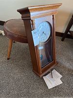 Wooden wall clock and round retro vinyl footstool on carpeted floor, clock leaning against stool.