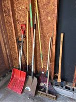 View of entire set of yard tools including shovels, rakes, broom, hoe, and axe, arranged standing in a corner.