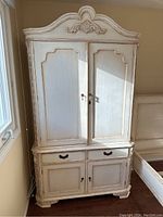 Front view of the French country antique style armoire showing decorative carved top, upper double cabinet doors, two drawers, and lower double cabinet doors.