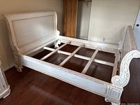 View of the queen bed frame fully assembled showing headboard, footboard, side rails, and wooden slats for mattress support. Painted in distressed white with carved decorative details.