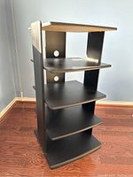 Side and angled front view of the dark grey/black media storage shelves on a wood floor showing multiple tiers and cable holes in the back.