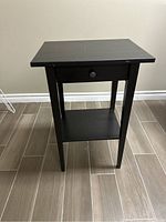 Front view of the black wood side table with drawer and shelf, showing minor scratches.