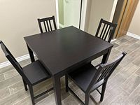 Square wooden dining table set with four matching wooden chairs arranged around it on tiled floor.