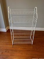 Full front view of white wired rack showing four shelves and metal frame standing on wooden floor against a light blue wall.
