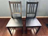 Two black wooden dining chairs with vertical slat backs and flat seats, placed on a wood floor against a light blue wall.