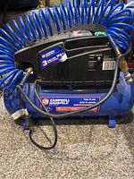 Front view showing the air compressor tank, motor housing, blue coiled air hose, and attached air gun tool.