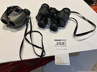 Two pairs of binoculars on a white surface with their neck straps visible. Tag labeled 'Binoculars' present.
