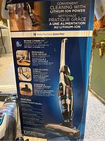 Close-up of vacuum box showing product features: 16V max lithium-ion powered suction, removable hand vacuum, 2-way folding handle, charging and storage station, multi-surface cleaning, and wash filter.