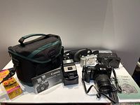 Overview photo showing Canon EOS 3000 camera with attached lens and strap, Lumix and Fujifilm cameras, Canon Speedlite 155A flash, camera bag, external flash accessory, and Kodak film roll.