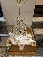 Photo of small chandelier placed inside cardboard box with protective tissue paper, showing hanging Swarovski crystals and gold-tone metal frame.