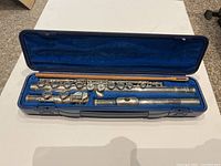 Open blue hard case showing the three-piece silver-tone Yamaha flute and wooden cleaning rod neatly placed inside blue velvet-lined compartments.