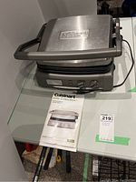 Full view of Cuisinart Elite Collection Griddler Deluxe appliance with instruction booklet on glass table surface.