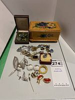 Overall view of jewelry lot on white surface showing rings, pins, clip-on earrings, and small wooden jewelry box with floral design.