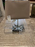 Schonbek lamp with curved crystal base, light grey rectangular fabric shade, and small square glass clock placed in front on floor carpet.