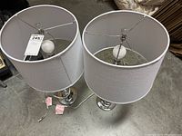 Two matching table lamps shown from above, displaying white drum shades and bulbs inside. Both lamps have cords visible with attached tags.