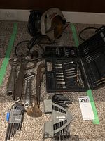 Photo showing Craftsman circular saw, wired and with rusted blade, multiple wrenches of different sizes, drill bit set case, and hex key sets arranged on carpet.