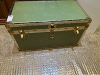 Full view of a rectangular light green trunk on carpet floor showing metal edged corners and brass locks.