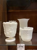 Four white milk glass style vases, differing in size and pattern, displayed on a shelf.