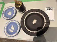 Four decorative Wedgwood pieces on table: two blue Jasperware plates with white relief and one black Jasperware plate with white relief along with black Jasperware table lighter