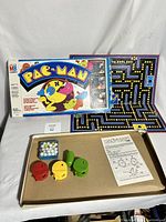 Pac-Man game box and unfolded game board with game pieces including yellow Pac-Man and three ghosts, white and blue marbles, instruction sheet