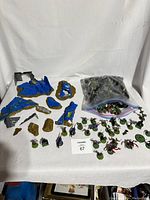 Overview of the table showing a large collection of painted fantasy miniatures mostly on green round bases, a ziplock bag of additional miniatures, and several painted plastic terrain pieces shaped as ruined blue stone structures and rocks.