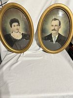 Two framed antique oval portraits side by side on a white cloth background showing husband and wife images behind convex bubble glass in gold frames.