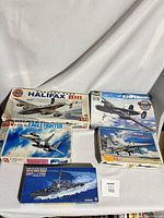 Five military model kits laid out on white cloth. Kits include multiple WWII and modern military aircraft and one naval ship. Boxes show visible wear and tear.