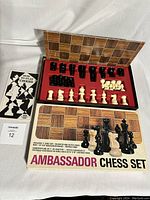 Photo showing the full set: black and white weighted, felted chess pieces arranged in a plastic insert, the folded 14x14 inch woodgrain chessboard behind, and the original box labeled 'Ambassador Chess Set'. Also includes a How to Play Chess instruction booklet.