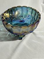 Blue iridescent carnival glass bowl with scalloped rim and embossed grape and floral pattern, shown from the side with three feet visible.