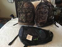 Three backpacks on a white surface: camouflage backpack front and center, gray and black patterned backpack next to it, and a black backpack with green logo lying in front.