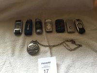 Six decorative lighters lined up with a silver-tone pocket watch with chain in front, placed on a soft beige fabric surface.