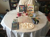 Wide shot showing full assortment of craft supplies on round table including kits, palette, plastic storage box, plush toy, and various art items.