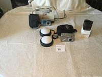 Photo showing two digital cameras, two dome-shaped light accessories, carrying cases, and a Budweiser portable speaker on a light fabric surface.