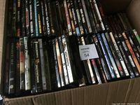 Photo showing assorted used DVD movies in two rows inside a cardboard box, titles visible include Olympus Has Fallen, The Biggest Loser, and various others.