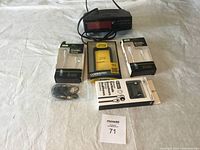 Photo showing OtterBox Commuter case in the center, two USB-C to Lightning charging cords on sides, a pair of wired earbuds in packaging, some unpackaged wired earbuds, and a black digital clock radio with red LED display at the top-center back.