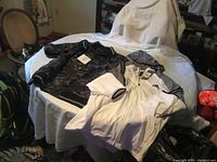 Two men's Adidas jackets laid on a white sheet on a bed, one white and gray with Adidas logo, the other black and gray camouflage design.