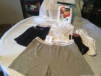 Photo showing the six items included in the lot all laid out on white fabric background. Items: gray and black Tommy Hilfiger boxer briefs, white t-shirt, 6-pack white tank tops in packaging, two pairs of white CCM socks (one pair still in package).