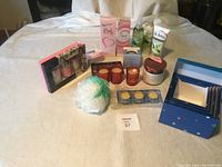 Assorted new beauty items on a white bed including skincare products, candles, makeup palette, bath sponge, and nested storage boxes