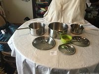 Three stainless steel pots with handles, three matching stainless steel lids, and a green silicone kitchen item arranged on a table.