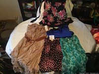 Photo showing five women's clothing items laid on bed including beige long dress with brown belt, blue dress/top, black floral top with pink flowers, black dress/skirt with pink heart pattern, turquoise floral garment.