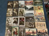 Top-down view of 24 vintage postcards arranged in rows showing front images with various scenes and subjects, including children, animals, flowers, ships, and scenic views.