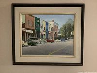 Framed oil painting showing street scene with colorful buildings, cars and pedestrians