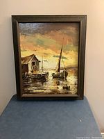 Frontal view of framed oil painting depicting two boats docked by a wooden shack at sunset
