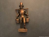 Front view of bronze/brass figurine labeled Hummel, showing man with top hat carrying two buckets on shoulder yoke