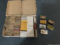 Two boxes filled with a large quantity of vintage matchbooks and matchboxes arranged with some boxed sets on the right side showing labels.