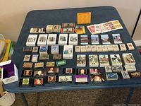 Overview of approximately 60 empty matchboxes and matchbooks displayed on a table with various vintage and travel-themed designs